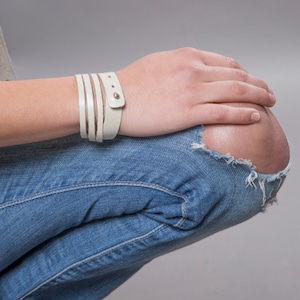 May include: A white leather bracelet with gold accents. The bracelet is wrapped around a person's wrist.