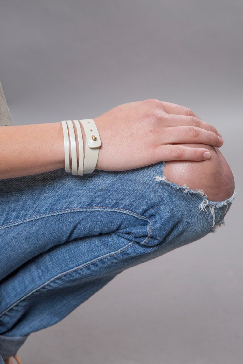 May include: A white leather bracelet with gold accents. The bracelet is wrapped around a person's wrist.