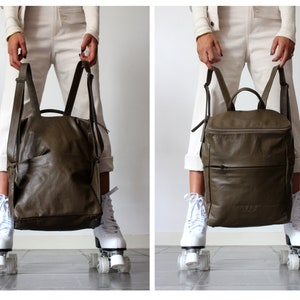 May include: A dark green leather backpack with a zippered pocket on the front. The backpack is being held by a person wearing white pants and white roller skates.
