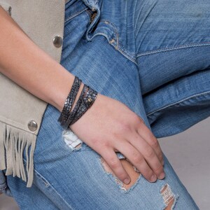 May include: A close-up of a person wearing a light brown suede waistcoat with fringe and blue jeans with distressed details. The person is wearing a black leather wrap bracelet with a gold accent.