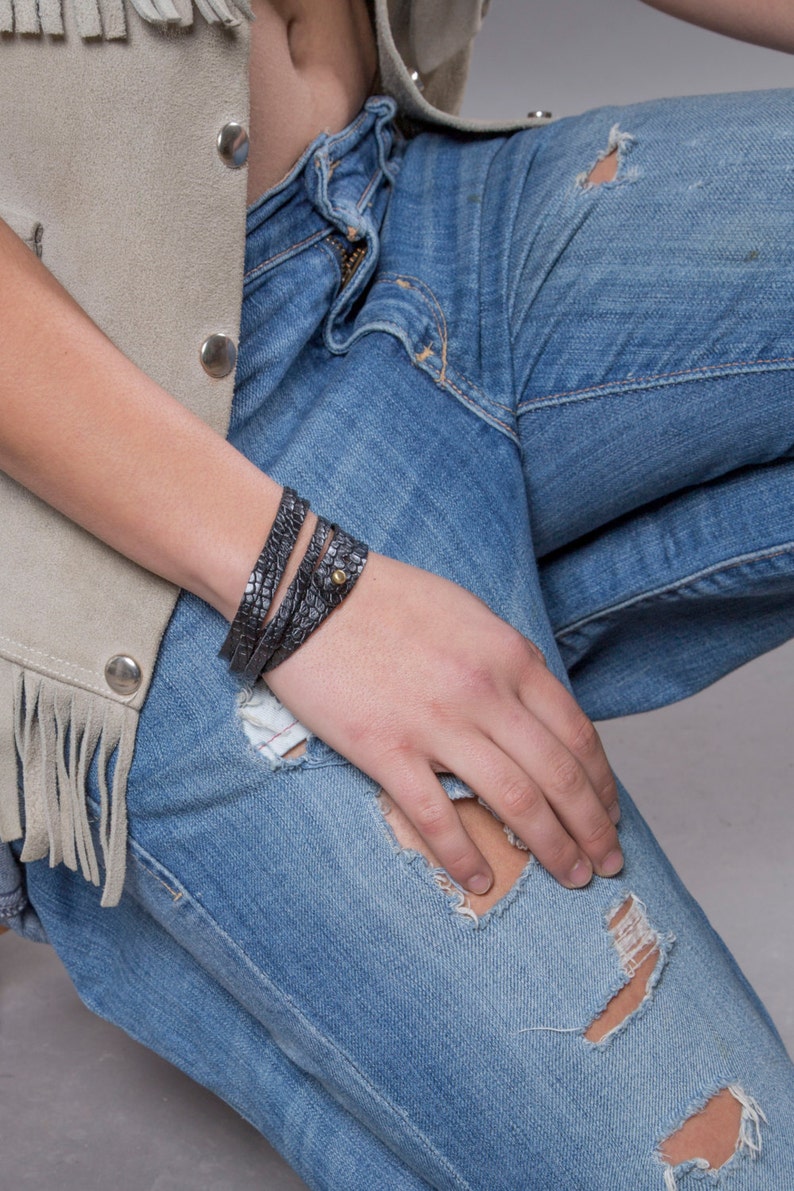 May include: A close-up of a person wearing a light brown suede waistcoat with fringe and blue jeans with distressed details. The person is wearing a black leather wrap bracelet with a gold accent.