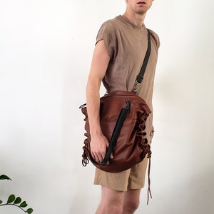 May include: A brown leather bag with a black adjustable strap. The bag has a gathered, ruffled design and is worn over the shoulder.