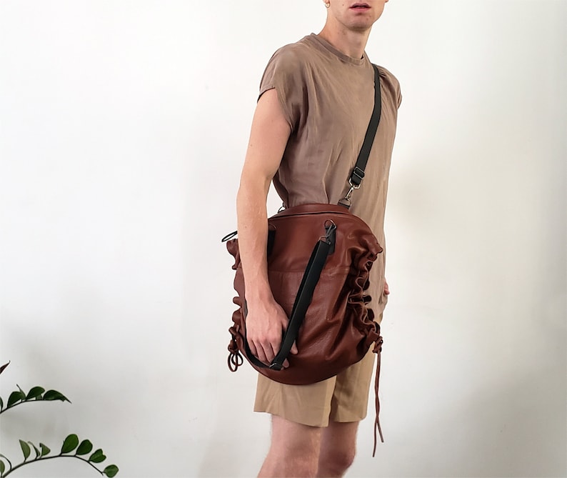 May include: A brown leather bag with a black adjustable strap. The bag has a gathered, ruffled design and is worn over the shoulder.