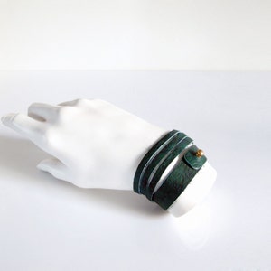 May include: A green leather wrap bracelet with a gold accent. The bracelet is wrapped around a white mannequin hand.