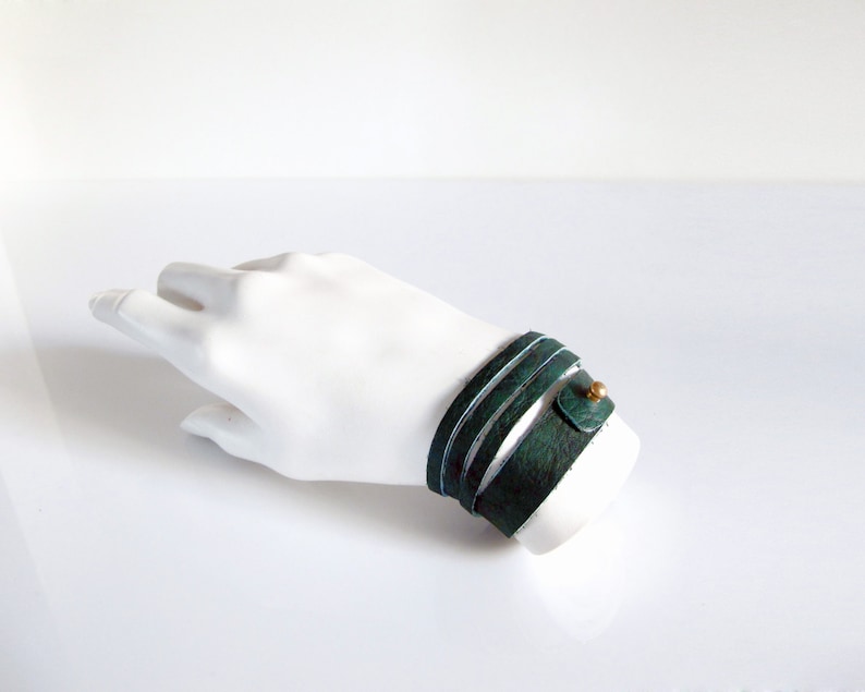 May include: A green leather wrap bracelet with a gold accent. The bracelet is wrapped around a white mannequin hand.