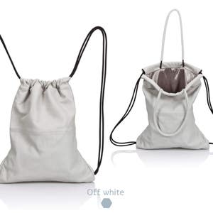 May include: Off-white drawstring backpack with black drawstrings. The bag is shown in two views: closed and open, revealing a silver interior. The bag has a simple, minimalist design, suitable for various uses.
