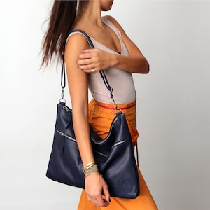 May include: A navy blue leather shoulder bag with a zipper closure and a long strap. The bag is worn by a person wearing a light pink tank top and an orange skirt.