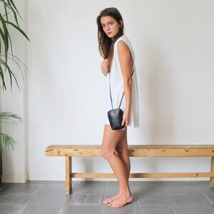 May include: A woman wearing a white sleeveless dress and a black crossbody bag. She is standing in front of a wooden bench.