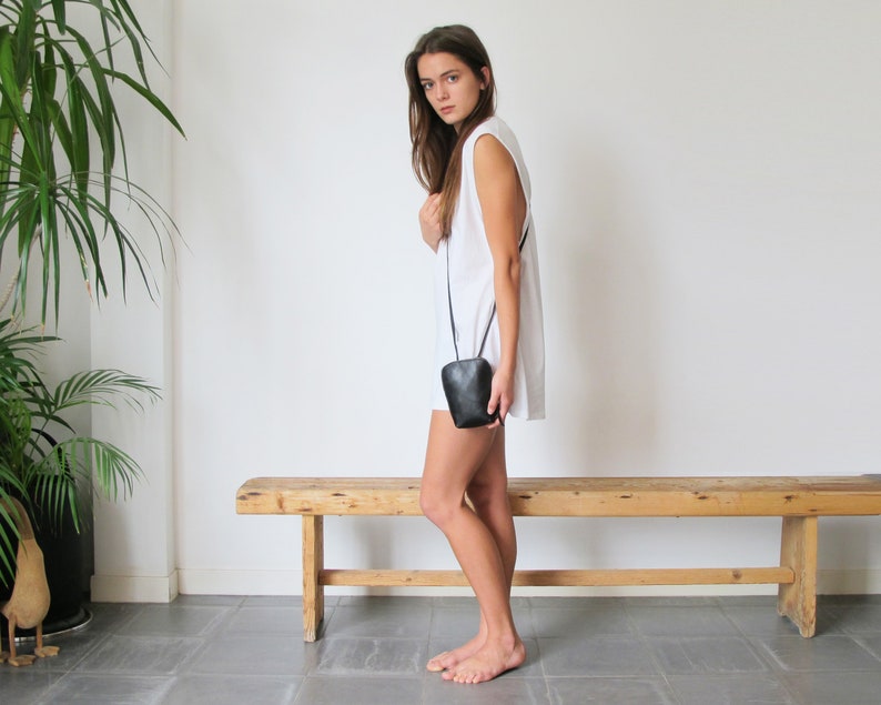 May include: A woman wearing a white sleeveless dress and a black crossbody bag. She is standing in front of a wooden bench.