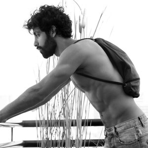 May include: A black and white photo of a person with a black backpack. The person is shirtless and wearing jeans. The person is leaning against a railing with their arm extended.