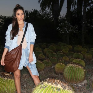 May include: A woman wearing a light blue denim jacket with a brown leather shoulder bag. She is standing in a field of cacti.
