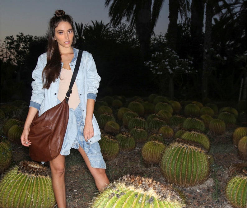 May include: A woman wearing a light blue denim jacket with a brown leather shoulder bag. She is standing in a field of cacti.