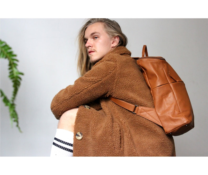 May include: A brown leather backpack with adjustable straps. The backpack is worn by a person wearing a brown fuzzy coat.