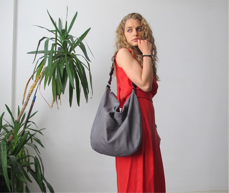 May include: A woman wearing a red dress and a grey leather shoulder bag. The bag has a long strap and is worn over the shoulder.