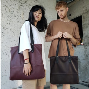 May include: Two people are holding leather tote bags. The woman is holding a burgundy bag with black straps. The man is holding a brown bag with black straps.