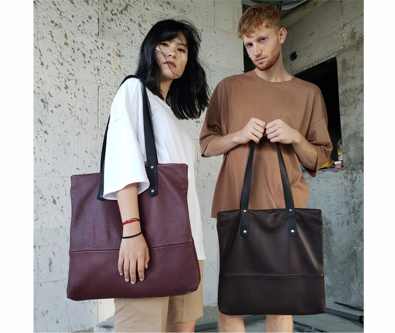 May include: Two people are holding leather tote bags. The woman is holding a burgundy bag with black straps. The man is holding a brown bag with black straps.