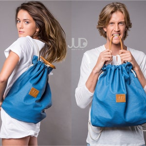 Puede incluir: Dos personas modelan una mochila azul de cordón con un parche de cuero marrón que dice "JUD". La mochila tiene un asa de madera y cordones blancos.