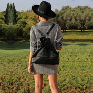 May include: A woman wearing a gray sparkly sweater and a black fedora hat is walking in a grassy field. She is carrying a black quilted backpack with a drawstring closure.