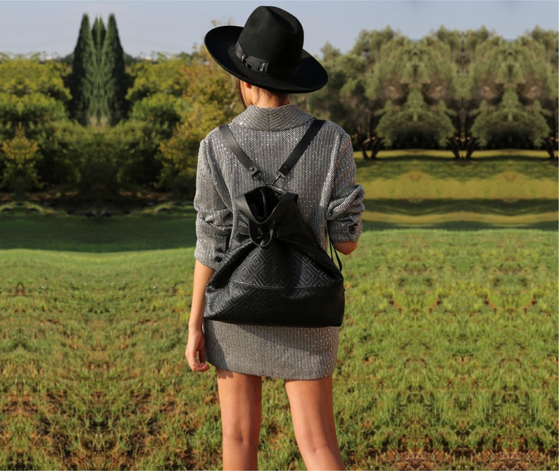 May include: A woman wearing a silver sparkly dress and a black fedora hat is walking in a grassy field. She is carrying a black leather backpack with a geometric pattern.