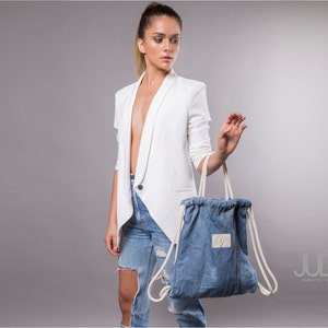 Może przedstawiać: Kobieta w białym blazerze i zniszczonych dżinsach trzyma niebieską torbę z sznurkiem z białych sznurków. Torba ma białą etykietę z napisem "JUD Made in NYC".