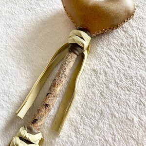 May include: A heart-shaped object with a smooth brown surface attached to a textured wooden stick. The connection point and the base of the stick are wrapped in cream-coloured leather strips that hang down as decorative ties along the handle.