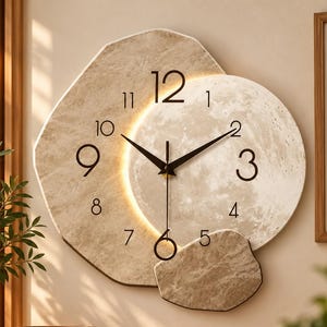 Puede incluir: Un reloj de pared con diseño en capas, que presenta una base grande e irregular con textura de piedra beige y una esfera circular similar a la luna. Incluye manecillas negras, números negros y una luz cálida integrada.