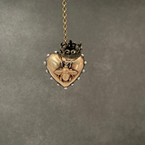 Puede incluir: Un colgante de coraz&oacute;n dorado con un dise&ntilde;o de abeja y una corona en la parte superior. El colgante cuelga de una cadena dorada.