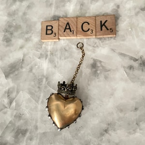 Puede incluir: Un colgante de coraz&oacute;n dorado con una corona en la parte superior, que cuelga de una cadena dorada. El colgante est&aacute; sobre un fondo blanco con la palabra "BACK" escrita en fichas de Scrabble.