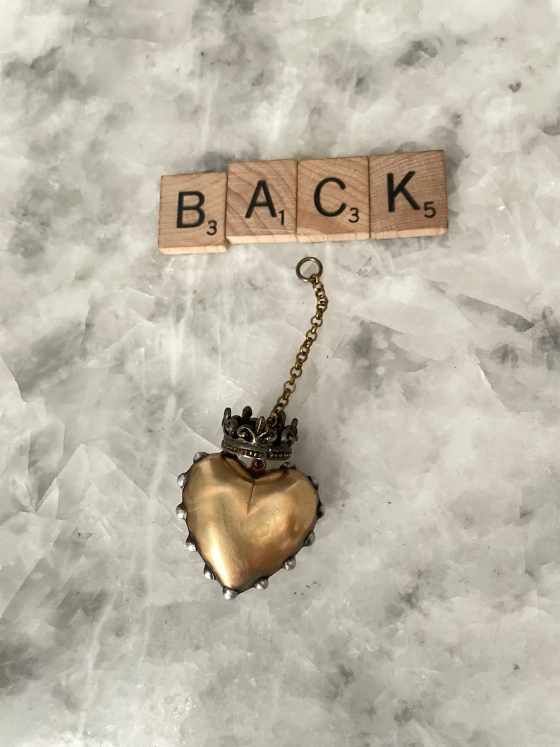 Puede incluir: Un colgante de coraz&oacute;n dorado con una corona en la parte superior, que cuelga de una cadena dorada. El colgante est&aacute; sobre un fondo blanco con la palabra "BACK" escrita en fichas de Scrabble.
