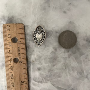 May include: A silver charm in the shape of a heart with a decorative border. The charm is about the size of a US quarter.