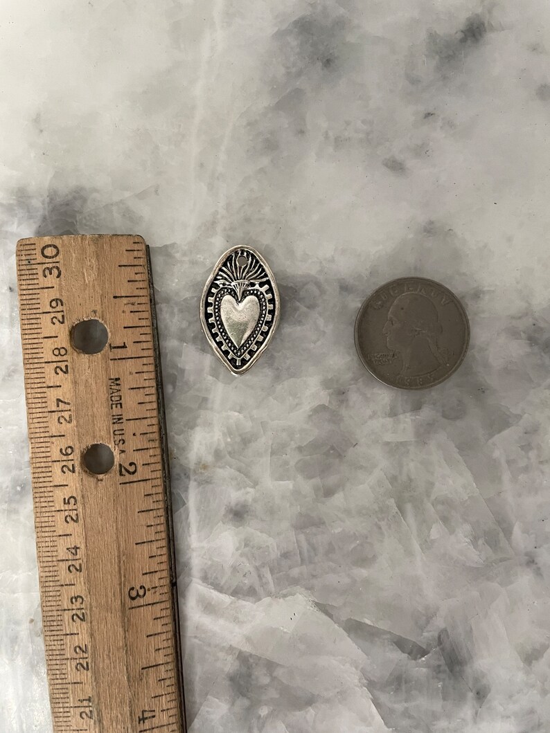 May include: A silver charm in the shape of a heart with a decorative border. The charm is about the size of a US quarter.