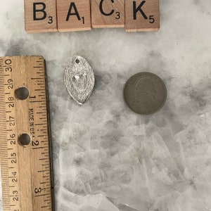 May include: A silver charm in the shape of a leaf with a textured surface. The charm is next to a ruler and a US quarter. The charm is approximately 1 inch in length. The letters B, A, C, and K are visible on the tiles above the charm.
