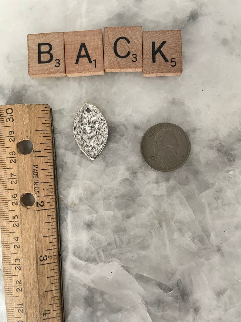 May include: A silver charm in the shape of a leaf with a textured surface. The charm is next to a ruler and a US quarter. The charm is approximately 1 inch in length. The letters B, A, C, and K are visible on the tiles above the charm.