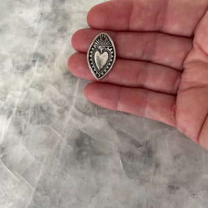 May include: A silver charm in the shape of an oval with a heart design inside. The heart has a raised design and a small flame at the top.