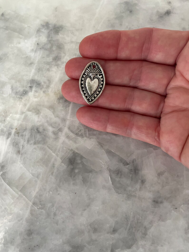 May include: A silver charm in the shape of an oval with a heart design inside. The heart has a raised design and a small flame at the top.