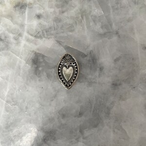 May include: A silver charm in the shape of a heart with a decorative border. The heart is surrounded by a leaf-shaped frame.
