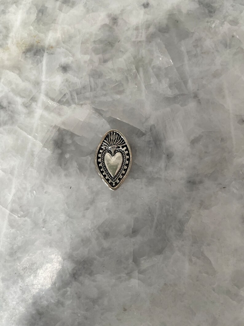 May include: A silver charm in the shape of a heart with a decorative border. The heart is surrounded by a leaf-shaped frame.