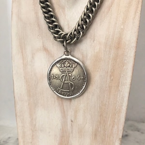 Könnte beinhalten: Eine silberne Kettenkette mit einem großen silbernen Anhänger mit einer Krone und der Inschrift "15.64".