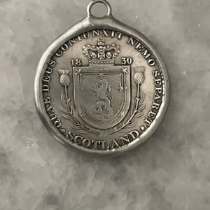 May include: Silver pendant featuring the Scottish coat of arms with a lion rampant, surrounded by the text "NEMO ME IMPUNE LACESSIT" and "1830", with the words "DUAL DEUS CONJUNXIT NEMO SEPARE" and "SCOTLAND" around the edge.