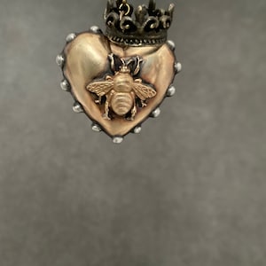 Puede incluir: Un colgante de coraz&oacute;n dorado con un dise&ntilde;o de abeja y una corona en la parte superior. El colgante cuelga de una cadena.
