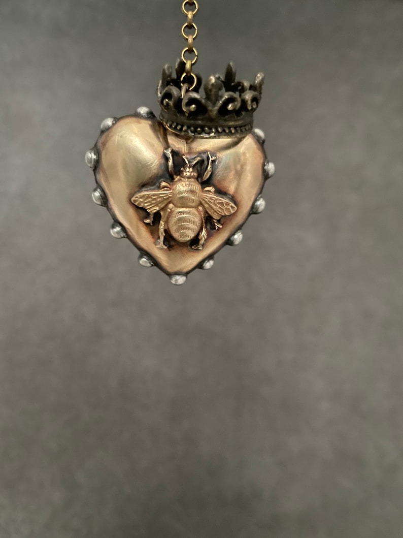 Puede incluir: Un colgante de coraz&oacute;n dorado con un dise&ntilde;o de abeja y una corona en la parte superior. El colgante cuelga de una cadena.