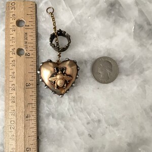 Puede incluir: Un colgante de coraz&oacute;n dorado con un dije de abeja y una cadena dorada con un anillo decorativo. El colgante tiene un di&aacute;metro de aproximadamente 5 cm.