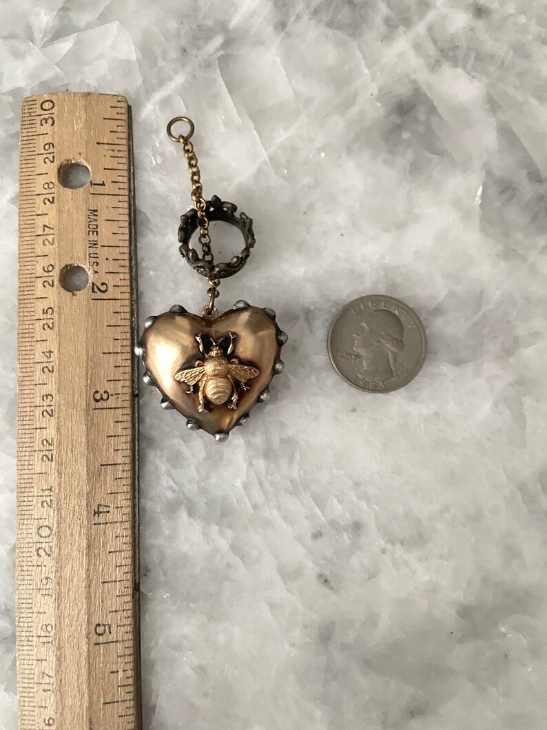 Puede incluir: Un colgante de coraz&oacute;n dorado con un dije de abeja y una cadena dorada con un anillo decorativo. El colgante tiene un di&aacute;metro de aproximadamente 5 cm.