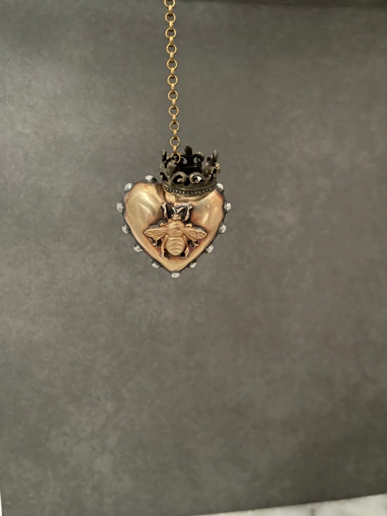 Puede incluir: Un colgante de coraz&oacute;n dorado con un dise&ntilde;o de abeja y una corona en la parte superior. El colgante cuelga de una cadena dorada.