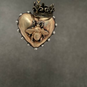 Puede incluir: Un colgante de coraz&oacute;n dorado con un dise&ntilde;o de abeja y una corona en la parte superior. El colgante cuelga de una cadena dorada.