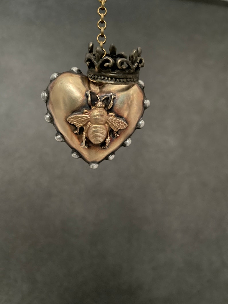 Puede incluir: Un colgante de coraz&oacute;n dorado con un dise&ntilde;o de abeja y una corona en la parte superior. El colgante cuelga de una cadena dorada.