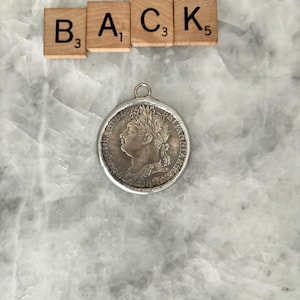 May include: Silver coin pendant with a profile of a woman with a crown on her head. The coin has the words "BRITANNIA" and "ONE PENNY" on it.