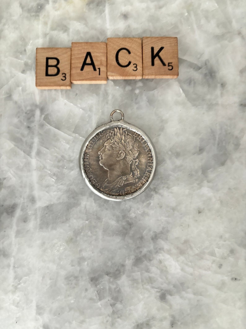 May include: Silver coin pendant with a profile of a woman with a crown on her head. The coin has the words "BRITANNIA" and "ONE PENNY" on it.