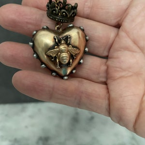 Puede incluir: Un colgante de coraz&oacute;n dorado con un dise&ntilde;o de abeja y un dije de corona en una cadena dorada. El colgante tiene una superficie texturizada y est&aacute; acentuado con tachuelas plateadas.