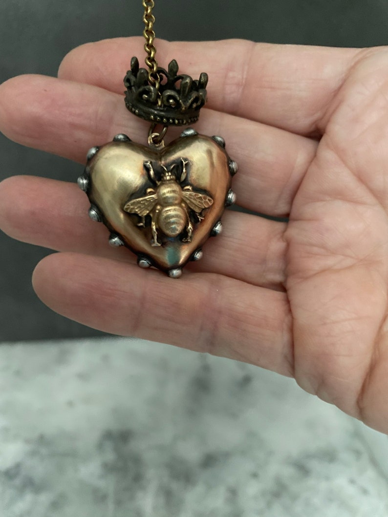 Puede incluir: Un colgante de coraz&oacute;n dorado con un dise&ntilde;o de abeja y un dije de corona en una cadena dorada. El colgante tiene una superficie texturizada y est&aacute; acentuado con tachuelas plateadas.